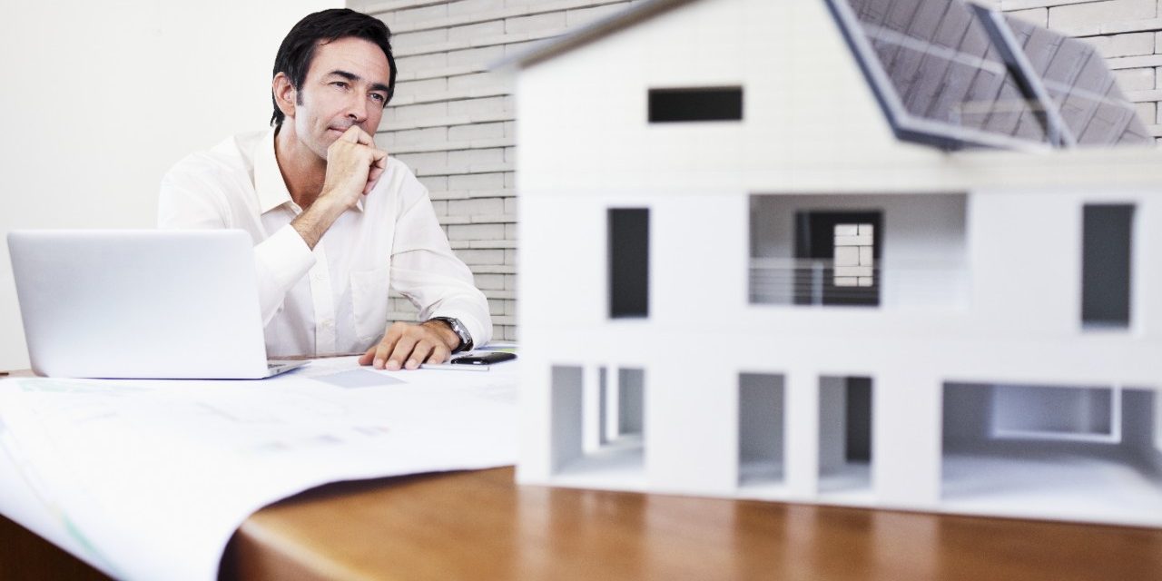 Architect sitting at office desk with blueprint and laptop looking at house mock-up 