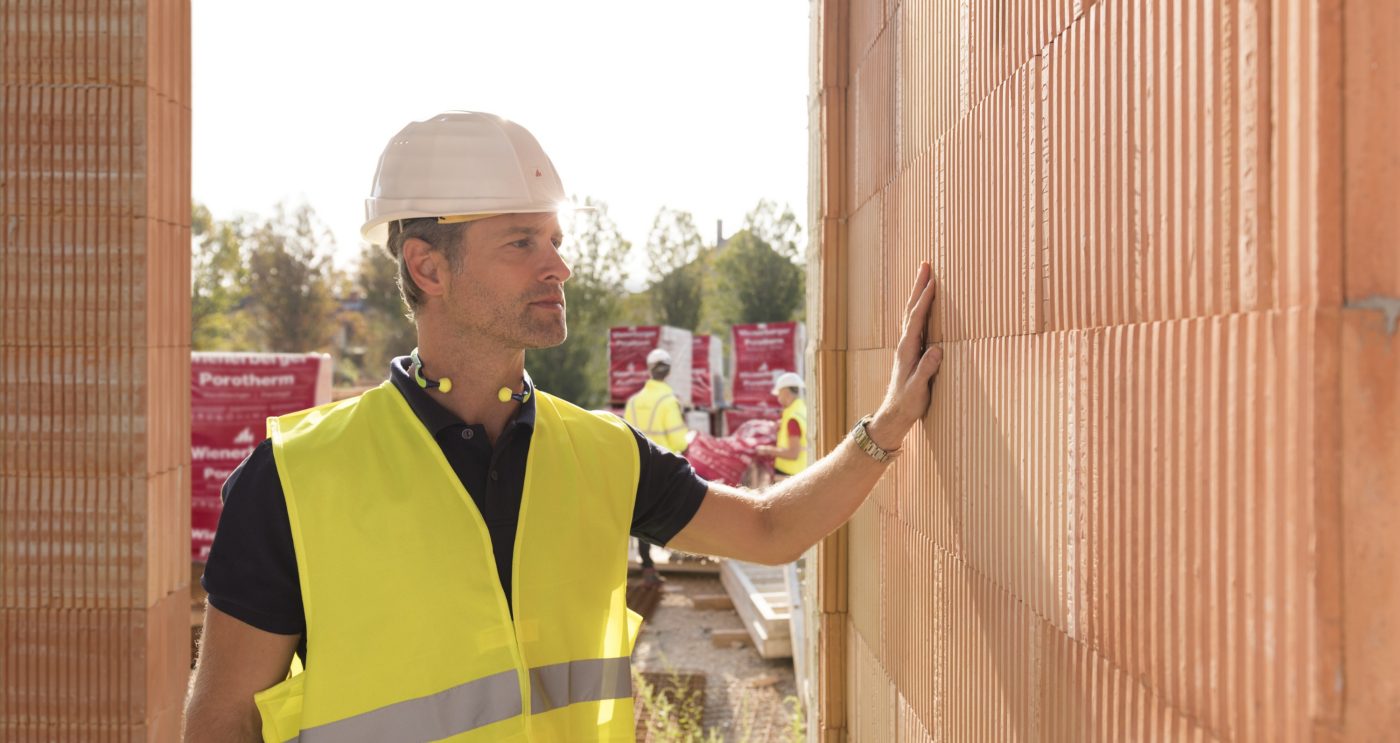 Builder on construction site touching clay block wall, construction workers and clay block pallets in the background