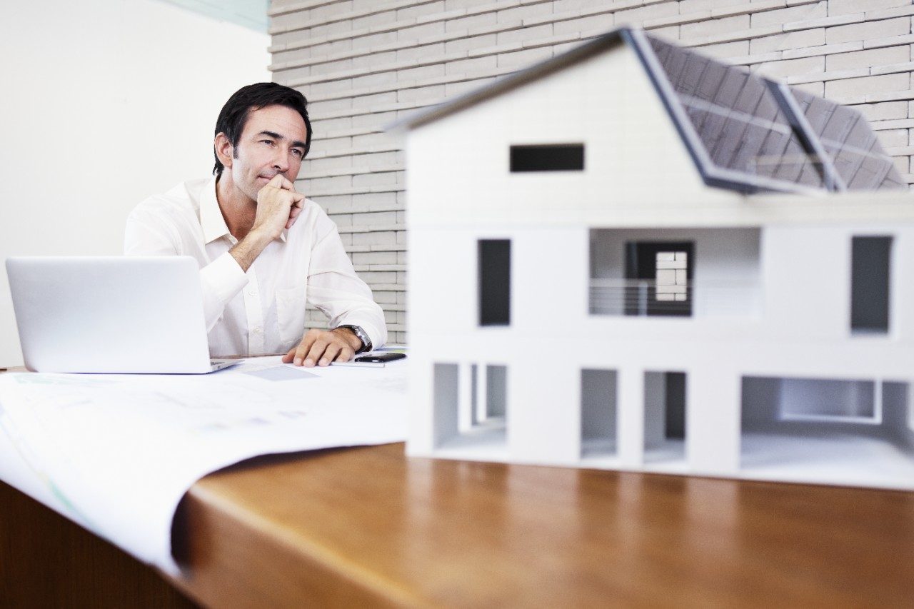 Architect sitting at office desk with blueprint and laptop looking at house mock-up 